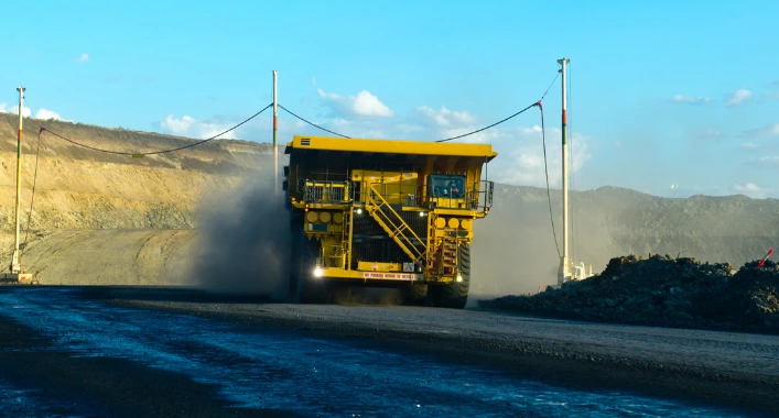 A haul truck moving slowly on a rough, uneven road, with dust clouds rising